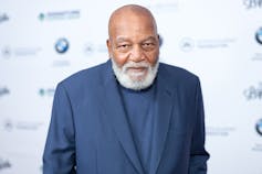 A black man with a grey beard wears a blue jacket as he stands in front of photographers.