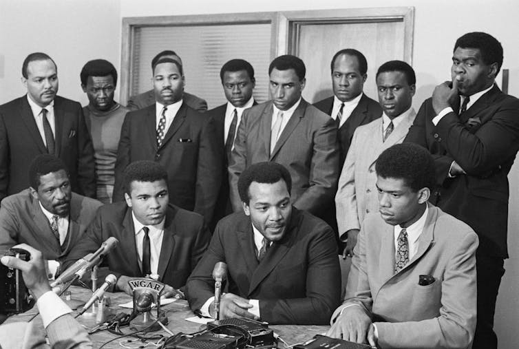 A group of professional Black athletes and politicians are gathered together during a meeting.