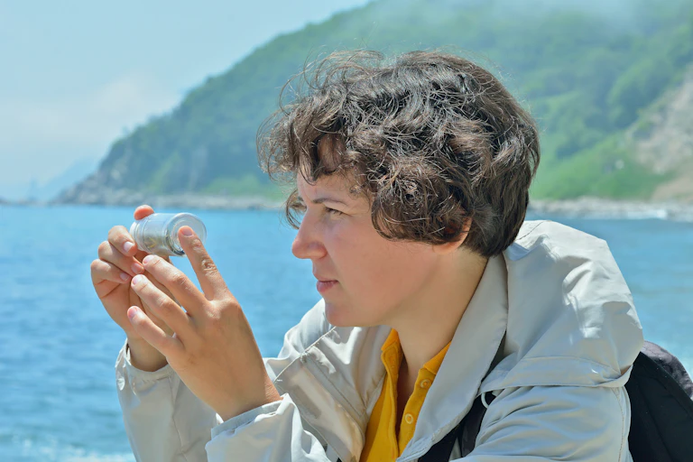 A woman in a parka next to a lake inspecting a jar with a sample up close