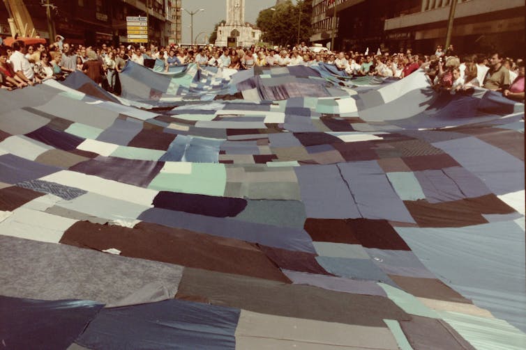 Decenas de personas sostienen una gran tela hecha con retazos de telas cosidos entre sí en una avenida principal de la ciudad de Bilbao.