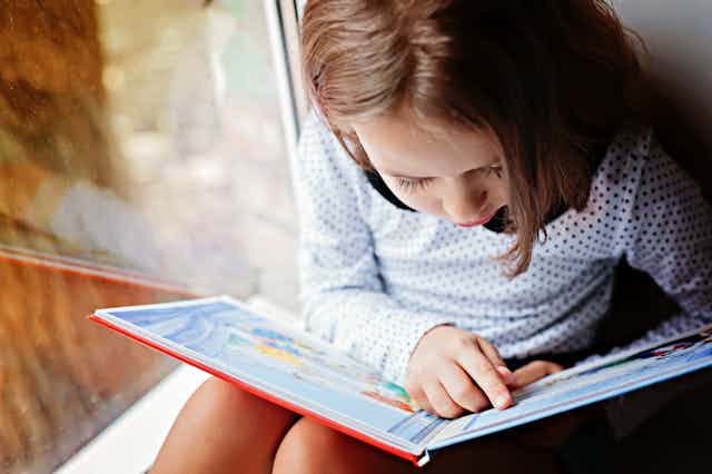 Una niña lee un libro al lado de una ventana