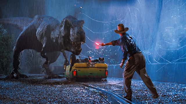 A T.rex stands over an upturned yellow car while a man in te foreground waves a bright red flare at it. 