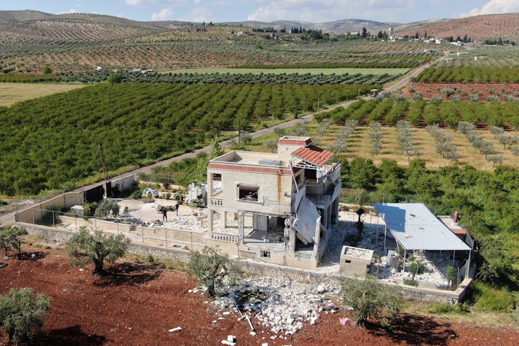 A large white building with rubble near it and farm fields behind it.
