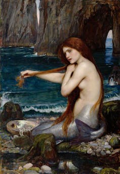 Una mujer con cola de pez está sentada en la orilla del mar en una playa mientras se peina el cabello largo.