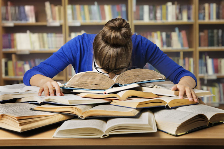 Una mujer hunde la nariz en un montón de libros, desesperada.