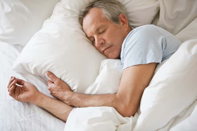 A Caucasian man, partially covered by a white blanket, sleeps in a bed.