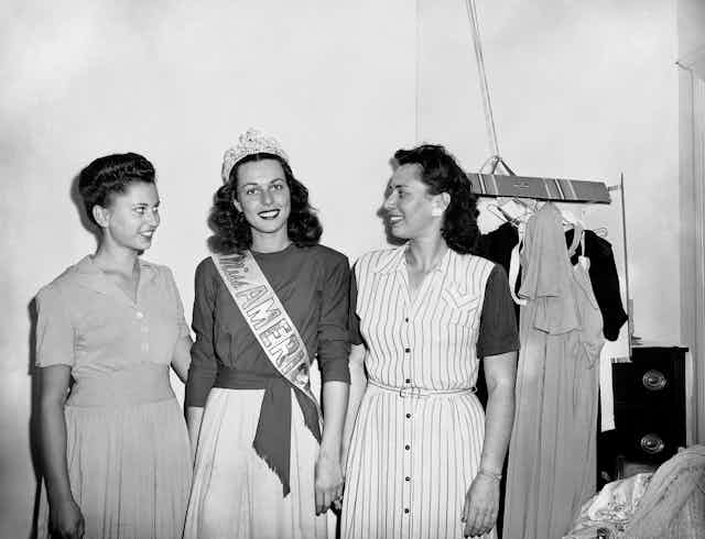 Three young, dark-haired women, one wearing a crown and a sash that says 'Miss America.'