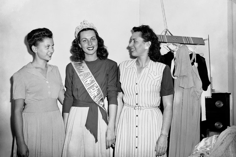 Three young, dark-haired women, one wearing a crown and a sash that says 'Miss America.'