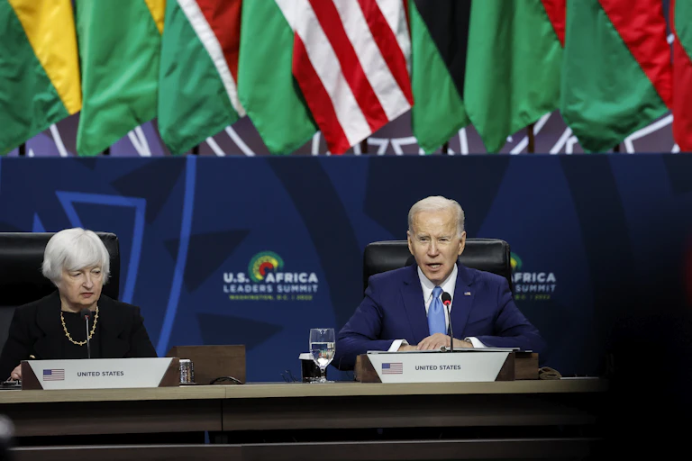 A gray-haired woman, wearing pearls, and a gray-haired, suited man at a stately table. Behind them hangs the American flag, surrounded by flags from numerous African countries.