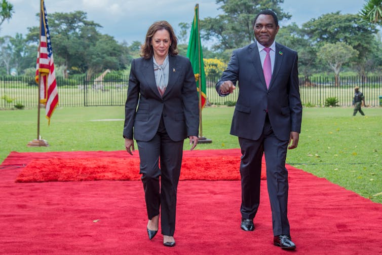 A woman in a pants suit on the left and suited man on the right walk on a red carpet. Behind them on the left stands the American flag. Behind them on the right stands the Zambian flag.