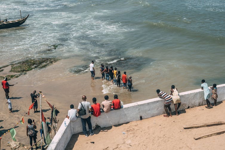 A group of people stand on sand, near an ocean both behind and in front of a long, elevated white wall.