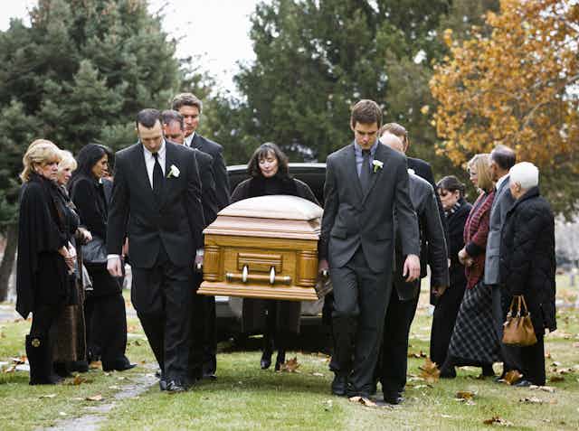 In a tree-lined cemetery, pallbearers dressed in black carry a coffin from the hearse to a gravesite.