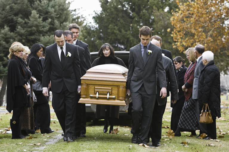 In a tree-lined cemetery, pallbearers dressed in black carry a coffin from the hearse to a gravesite.