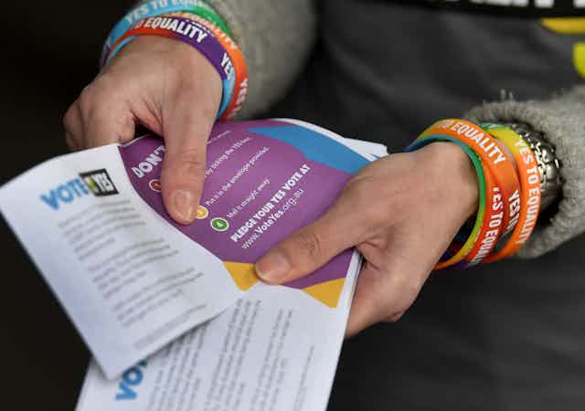 A person holds 'vote yes' pamphlets for the same sex marriage referendum. They wear colourful bracelets on their wrists that say 'marriage equality'.