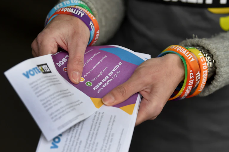 A person holds 'vote yes' pamphlets for the same sex marriage referendum. They wear colourful bracelets on their wrists that say 'marriage equality'.