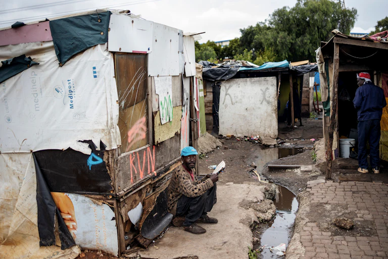 A man seated on the ground reads a magazine at an informal settlement