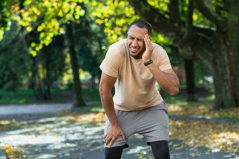 A man standing outside in a park holds his head in pain.