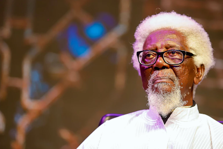 An elderly man with white hair and beard and a white robe and glasses sits looking straight ahead of him.