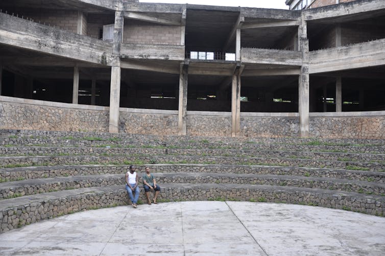 A man and a woman sit on stairs made from stone in an arena with a concrete building behind them.