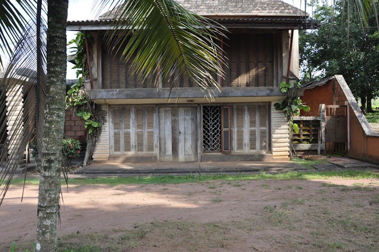 A two-storey house with large wooden doors and the entire front comprised of slatted wooden shutters in a yard with a palm tree.