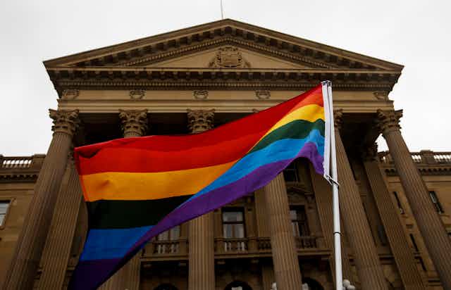 A large rainbow flag in front of a stone legislative building.
