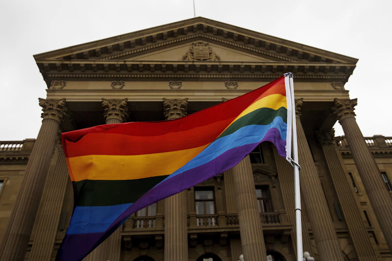 A large rainbow flag in front of a stone legislative building.