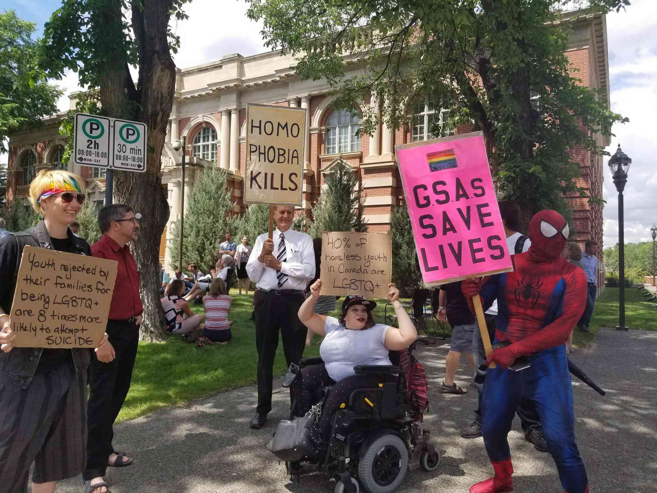 A group of people, including a person in a wheelchair, hold signs urging the protection of gay-straight alliances.