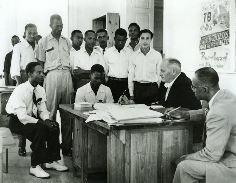 المجندون الكاريبيون وقفوا حول مكتب مع رجل أبيض يبحث في الأوراق.