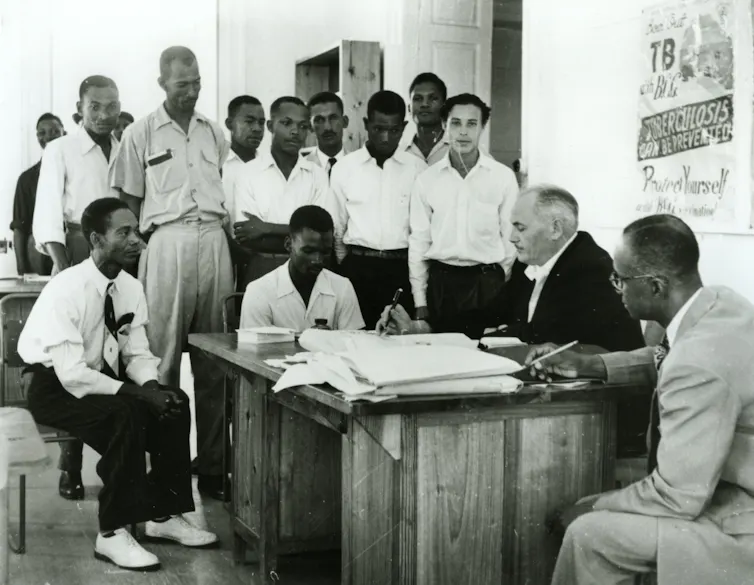 Caribbean recruits stood around an office with a white man looking through papers.