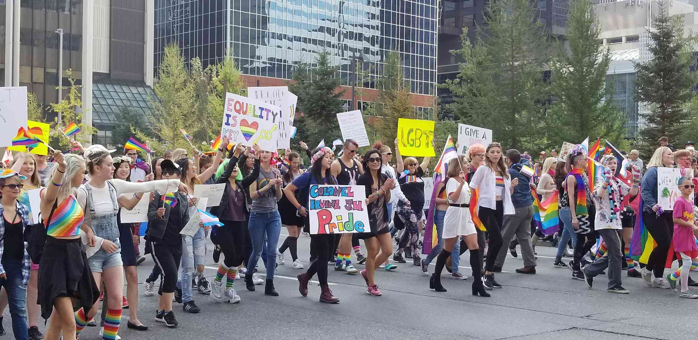 People march in a parade, some wearing rainbow clothing and carrying signs.