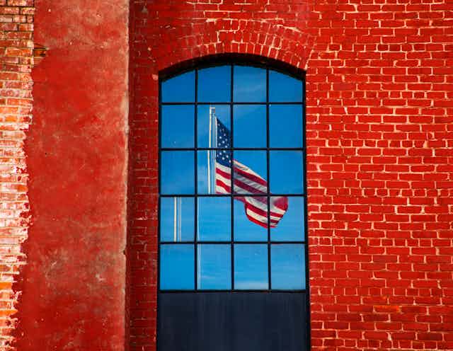 A brick wall with windows shows the reflection of a fractured American flag.