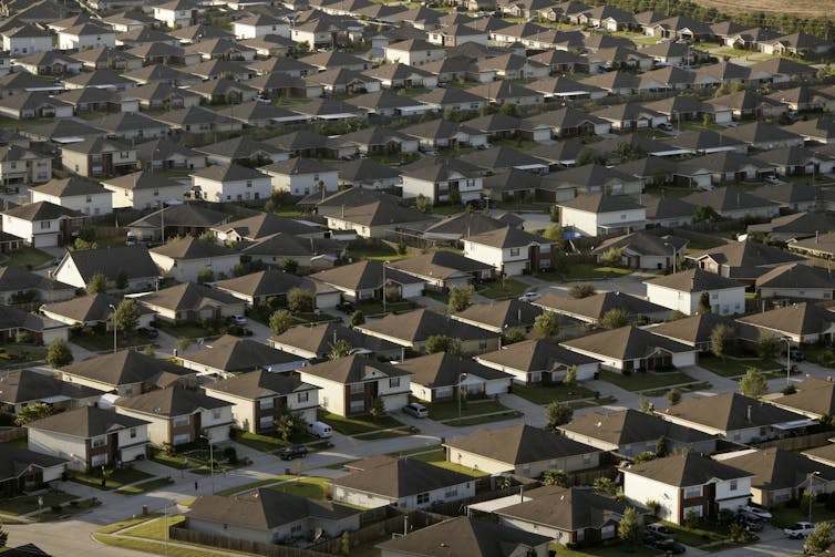 An aerial view shows suburban houses, all similar with dark roofs and white exteriors.