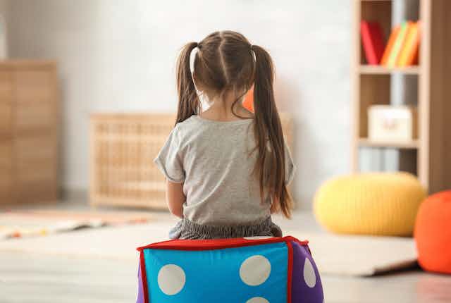 Una niña sentada en un cojín de espaldas a la cámara mira a la pared de una habitación con juguetes y muebles.