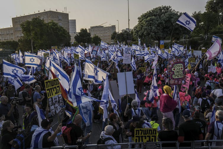 People at a protest wave blue and white Israeli flags featuring the Star of David. They also hold placards in Hebrew.