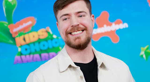 Young man with a beard and moustache grins against a brightly colored background.