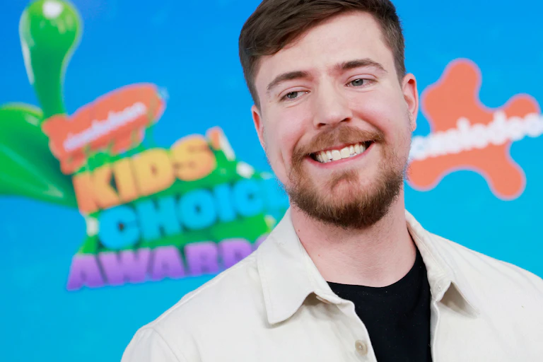 Young man with a beard and moustache grins against a brightly colored background.