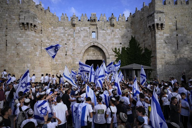 People at a march wave Israeli flags in front of an old wall with a gate.