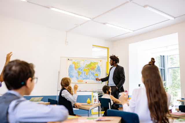 A teacher points at a world map, while students sitting at desk watch.