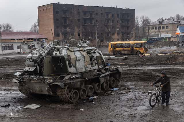 A man pushes his bike through mud and debris past a destroyed Russian tank in front of the central train station that was used as a Russian base on March 30, 2022 in Trostyanets, Ukraine
