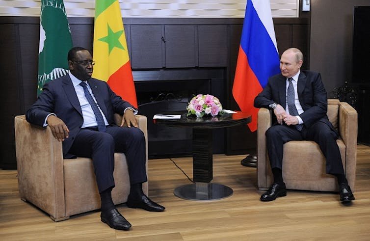 Two men talk while sitting on couches in front of the flags of Russia and Senegal.