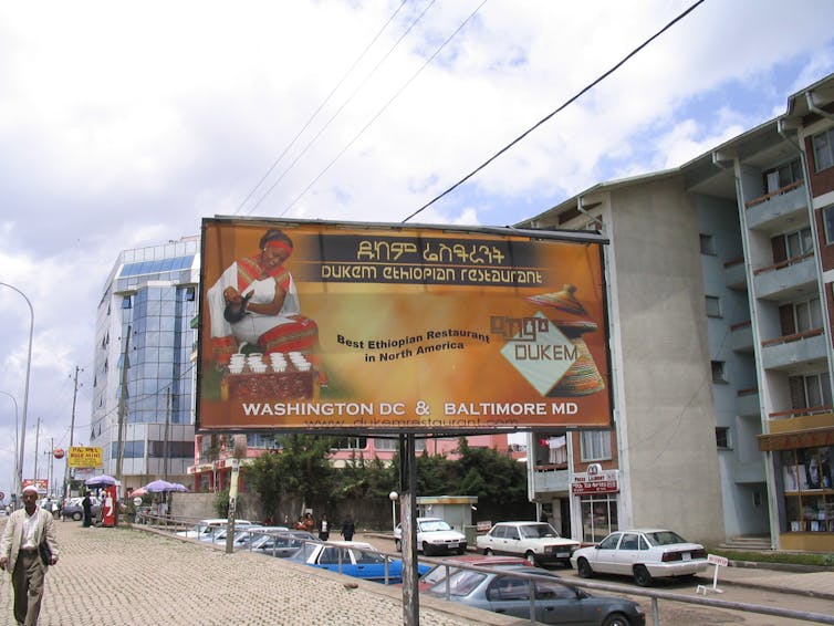 A main Addis Ababa street leading to the airport, Bole Road, with high-rise buildings and a billboard featuring a restaurant advert showing a woman in Ethiopian robes pouring coffee into rows of cups.