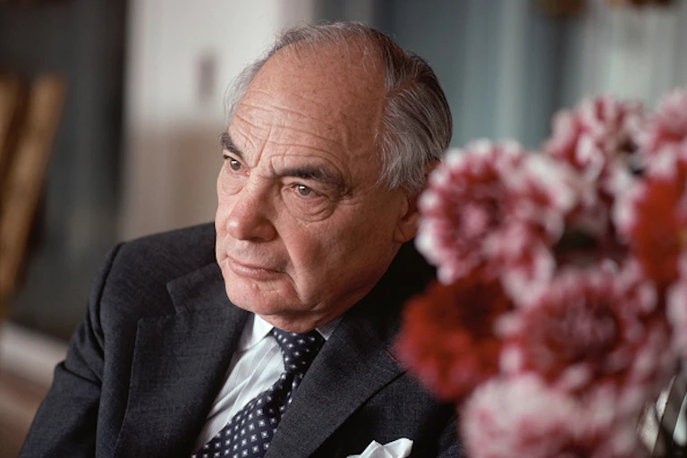 A serious looking man wearing a tie and suit tie sits at a table displaying a bunch of flowers.