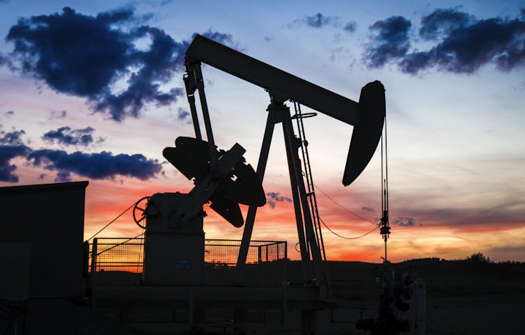 A large pumpjack resembling a tractor with a pick-axe head is seen against a blazing colourful sky.