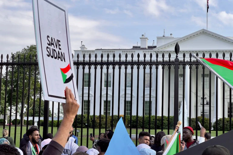A crowd of people holding flags stand in front of a large white building, with one placard held up that says 'save Sudan'