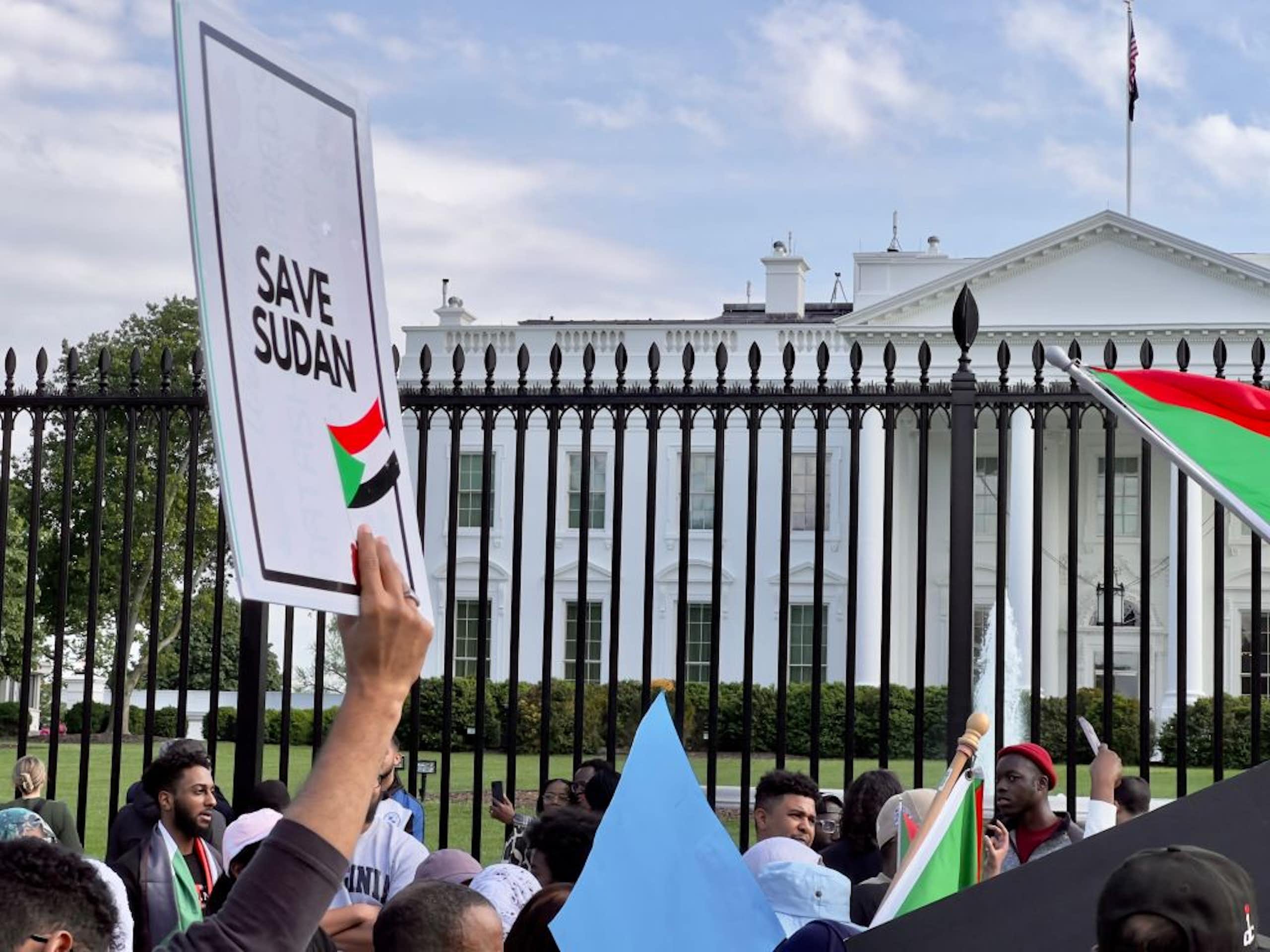 A crowd of people holding flags stand in front of a large white building, with one placard held up that says 'save Sudan'