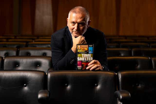 A man dressed in black sitting in an auditorium holding up his book, called Time Shelter.