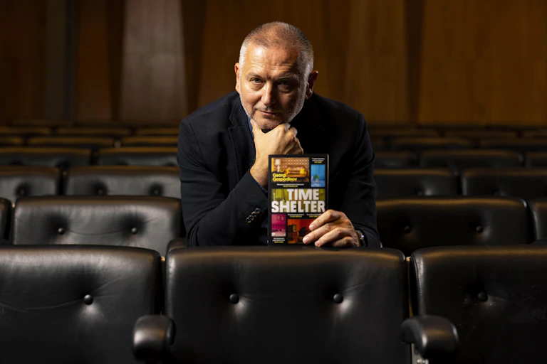 A man dressed in black sitting in an auditorium holding up his book, called Time Shelter.