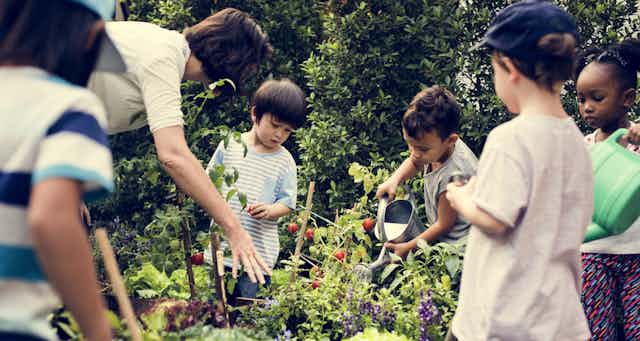 Children seen standing around a garden holding cans and touching plants.