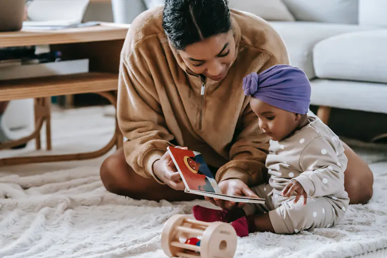 A mother seen on the floor with older baby and a board book.