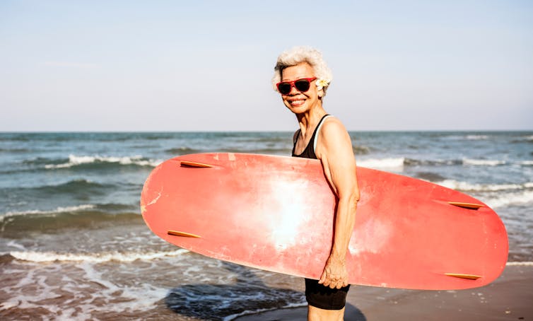 Woman with grey hair, sunglasses and surf board standing at the edge of the sea.